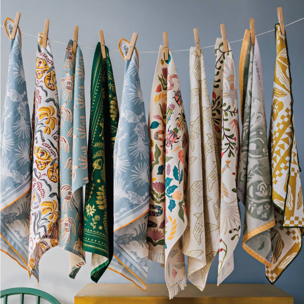 Colorful patterned tea towels hanging on a line against a gray wall with a yellow cabinet and green chair.